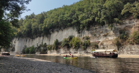 Riva fluviale vicino a Catus, Occitania, Francia, con persone in canoa e barca sotto scogliere alberate in una giornata di sole.