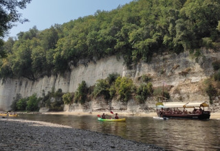 Riva fluviale vicino a Catus, Occitania, Francia, con persone in canoa e barca sotto scogliere alberate in una giornata di sole.