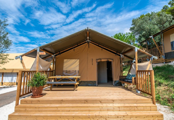 Safari teltet Lodge tent Bergerac Deluxe med træterrasse, picnicbord og bænk under en blå himmel.