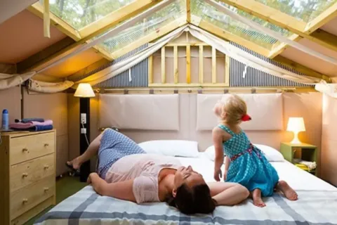 A woman and a child relax on a bed under a glass roof in Airlodge 1 at Chianti Glamping Resort, Italy.