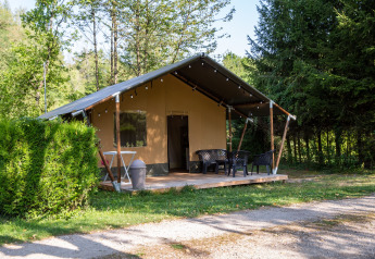 Safari-Zelt auf einem grünen, bewaldeten Campingplatz mit Terrasse und Gartenmöbeln im Freien.