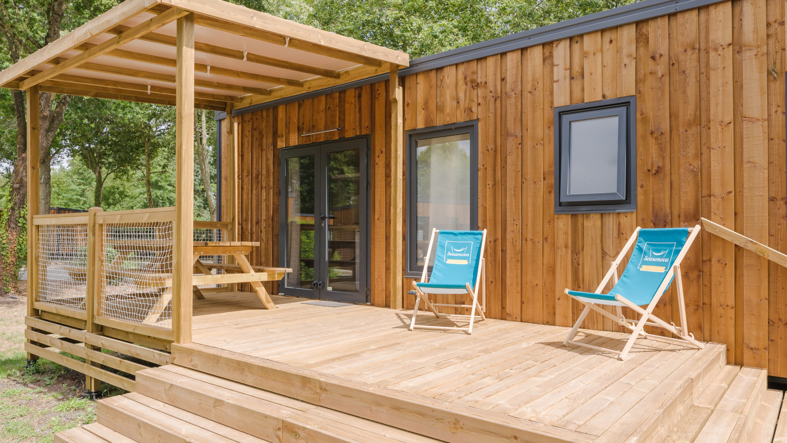 Houten lodge met overdekt terras, twee blauwe ligstoelen en picknicktafel in een natuurlijke omgeving.