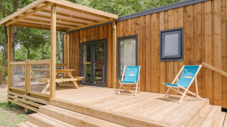 Casetta in legno con veranda coperta, due sdraio blu e tavolo da picnic immersi nel verde di un lodge.