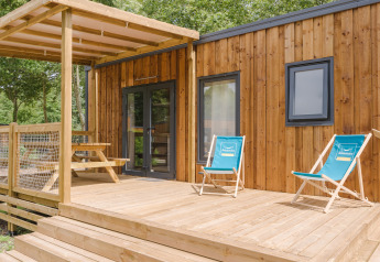 Casetta in legno con veranda coperta, due sdraio blu e tavolo da picnic immersi nel verde di un lodge.