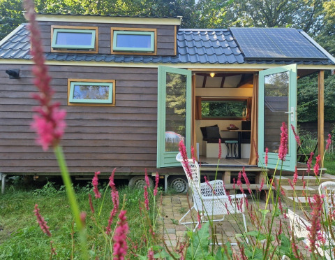 Tiny house Hans en Grietje, panneaux solaires, portes ouvertes, chaise de jardin et fleurs devant.