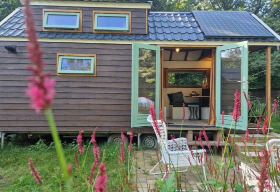 Tiny house Hans en Grietje, panneaux solaires, portes ouvertes, chaise de jardin et fleurs devant.