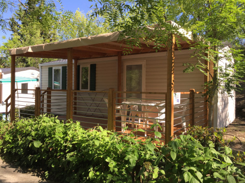 Mobil-home Royal avec terrasse couverte, entouré d’arbustes et d’arbres sous un ciel bleu clair.