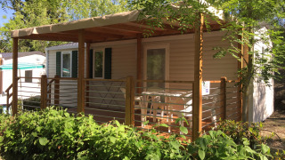 Mobil-home Royal avec terrasse couverte, entouré d’arbustes et d’arbres sous un ciel bleu clair.