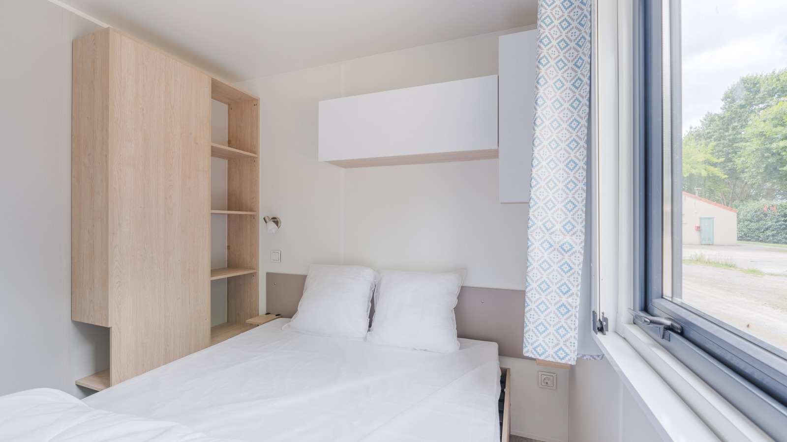 Lichte en moderne lodgekamer met tweepersoonsbed, open raam, kast en blauw patroon gordijn.