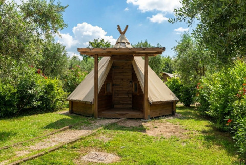 Tipi Junior tent omgeven door groen en bomen onder een blauwe lucht, ideaal voor een unieke kampeerervaring.