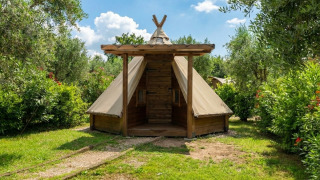 Tienda Tipi Junior ubicada entre jardines verdes y árboles, bajo un cielo azul, ideal para acampar cómodo.