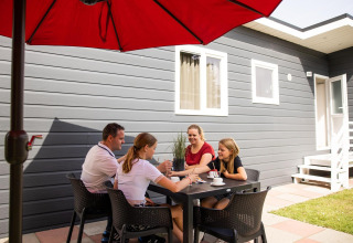 Familie sidder udenfor under en rød parasol ved et sort bord ved Group Yard Chalets på De Scherpenhof, NL.