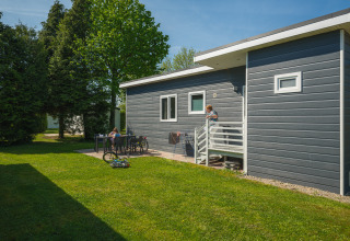 Moderne chalet i De Scherpenhof, Holland, med grøn have, cykler og personer der nyder udendørslivet.