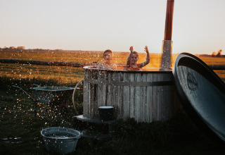 Dos niños juegan y salpican agua en una bañera al aire libre, con campos y atardecer de fondo.