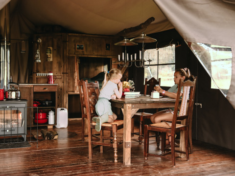 Two children enjoy a rustic glamping cabin, sitting at a wooden table surrounded by cozy wooden furniture.