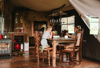 Two children enjoy a rustic glamping cabin, sitting at a wooden table surrounded by cozy wooden furniture.
