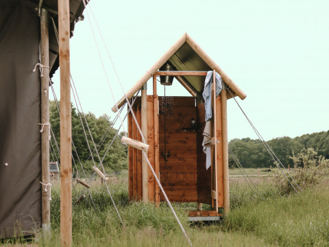 Wooden outdoor shower at a glamping site, surrounded by tents and greenery in a natural open setting.