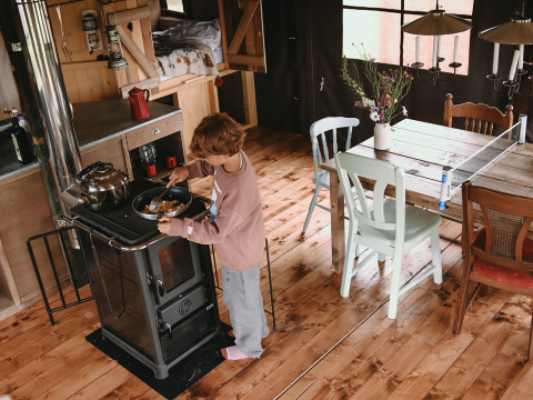 A child cooking at a stove in a cozy glamping cabin with rustic wooden floor and a dining table nearby.