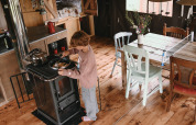 A child cooking at a stove in a cozy glamping cabin with rustic wooden floor and a dining table nearby.