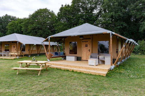 En moderne safaritelt med træveranda og udendørs siddeplads i naturskønne omgivelser nær skov.