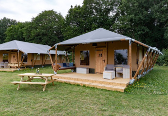 Modernes Safarizelt mit Holzveranda und Sitzbereich im Freien, gelegen an einer Wiese mit Bäumen.