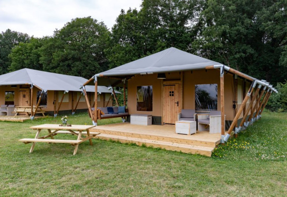 En moderne safaritelt med træveranda og udendørs siddeplads i naturskønne omgivelser nær skov.