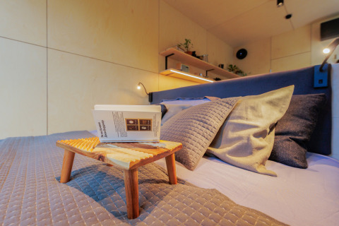 Chambre chaleureuse d’une tiny house avec plateau en bois, coussins, étagère, livre et éclairage doux.