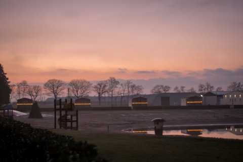 Tramonto su Mobi Lodge ad Arden Parks Tulderheyde in Belgio con casette illuminate e laghetto riflettente.