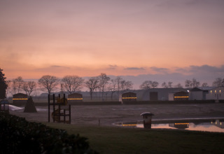 Tramonto su Mobi Lodge ad Arden Parks Tulderheyde in Belgio con casette illuminate e laghetto riflettente.