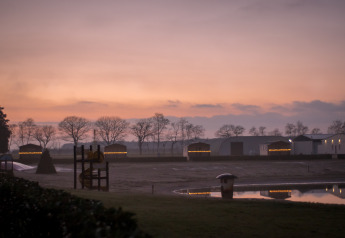 Tramonto su Mobi Lodge ad Arden Parks Tulderheyde in Belgio con casette illuminate e laghetto riflettente.