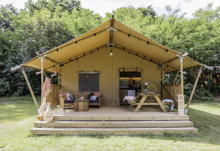 Tente safari avec terrasse en bois, coin salon extérieur et table, entourée d’arbres et de verdure.
