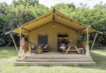Safari-Zelt mit Holzterrasse, Outdoor-Sitzgelegenheiten und Tisch, umgeben von grünem Wald.