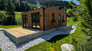 Cabane moderne en bois Arlberg avec grandes fenêtres, entourée de verdure, pierres et montagne.