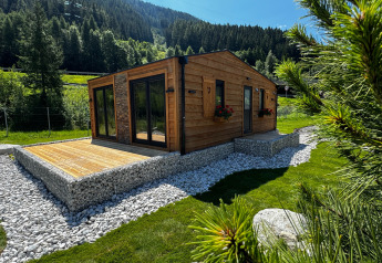 Cabaña Arlberg de madera moderna con terraza, rodeada de árboles, césped verde y muros de piedra.
