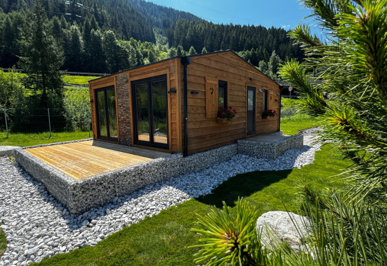 Cabane moderne en bois Arlberg avec grandes fenêtres, entourée de verdure, pierres et montagne.
