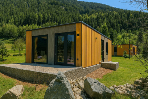 Moderne træhytte ved en lodge med terrasse og store vinduer, omgivet af grønne bakker og skov.