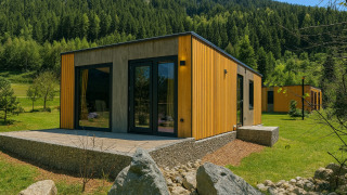 Modern wood cabin lodge with a terrace and large windows, surrounded by green hills and forest trees.