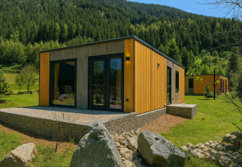 Lodge moderne en bois avec terrasse et grandes fenêtres, entourée de collines verdoyantes et de forêts.