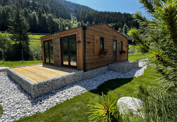 Cabaña de madera con terraza en Camping Arlberg, Austria, rodeada de colinas y árboles verdes.