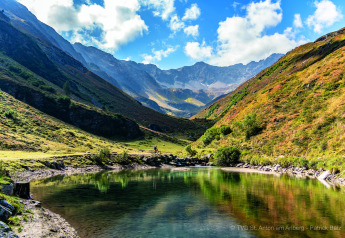 Paisaje montañoso con lago, colinas verdes, ciclista en sendero y cielos azules cerca de una cabaña alpina.