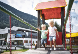 Twee jonge kinderen spelen op een speeltuinbrug bij een lodge, met bergen op de achtergrond.