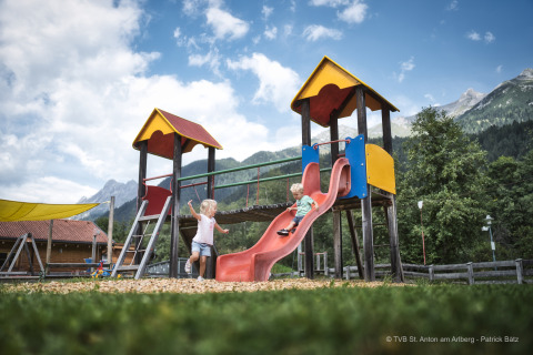 Deux enfants jouent sur un toboggan coloré près d’un chalet avec des montagnes en arrière-plan.