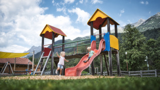 Deux enfants jouent sur un toboggan coloré près d’un chalet avec des montagnes en arrière-plan.