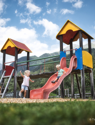 Due bambini giocano su uno scivolo colorato vicino a un rifugio, con le montagne sullo sfondo.