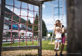 A woman helps a child play on gym rings at an outdoor playground near a mountain lodge on a sunny day.