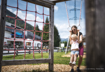 Una mujer ayuda a una niña a jugar en los aros de gimnasia de un parque infantil junto a una cabaña de montaña.