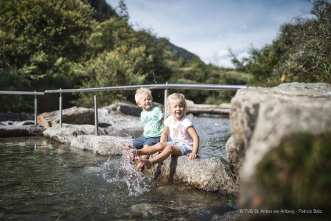 Deux enfants assis sur une pierre près d’un ruisseau jouent près d’un lodge en montagne par beau temps.