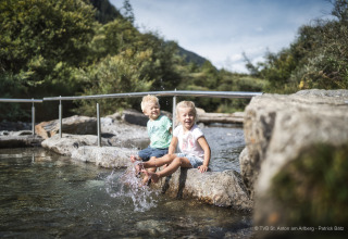 To børn sidder på en sten og plasker med fødderne i en bæk ved en lodge omgivet af bjerge og natur.
