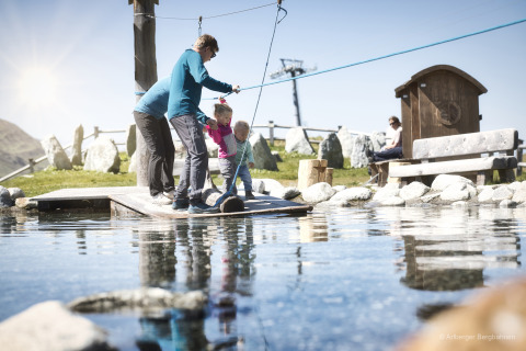 Family playing at a small pond by a lodge with mountain scenery in the background on a sunny day.