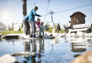 Family playing at a small pond by a lodge with mountain scenery in the background on a sunny day.
